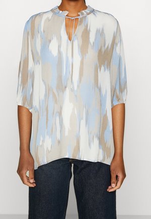 Woman wearing a loose blouse with blue, beige, and white abstract pattern, three-quarter sleeves, ruffled neckline, and dark jeans.