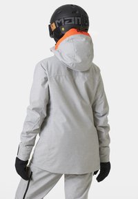 Šedá lyžařská bunda s kapucí, zdobená oranžovým akcentem, texturovaná tkanina a černé rukavice. Pro bezpečnost je na hlavě lyžařská přilba.
