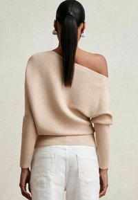 Beige gerippter Strickpullover mit Off-Shoulder-Design und langen engen Ärmeln, kombiniert mit weißen hoch taillierten Jeans.