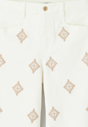 Pantalon blanc avec des motifs en losange brodés marron, espacés uniformément sur le tissu, et une fermeture boutonnée marron visible à la taille.
