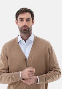 Beige Strickjacke aus weichem Material, mit V-Ausschnitt, Knopfleiste und gerippten Bündchen, getragen über einem hellblauen Hemd mit Kragen.
