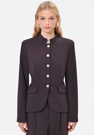 Donna bionda che indossa una giacca scura su misura con bottoni argentati e grandi orecchini dorati vistosi su sfondo bianco uniforme.