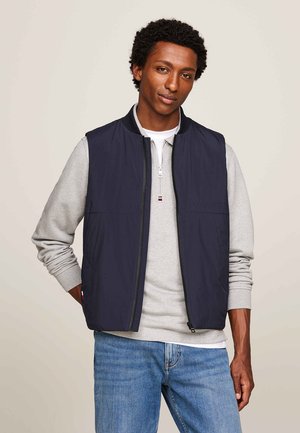 Marinefarbene ärmellose Weste mit hohem Kragen, Frontreißverschluss und Seitentaschen, kombiniert mit einem grauen Sweatshirt und blauen Jeans.