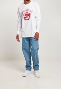 Henkilö seisoo päällä valkoinen pitkähiha-paita, jossa on punainen logo, siniset farkut ja valkoiset tennarit vaaleaa taustaa vasten.