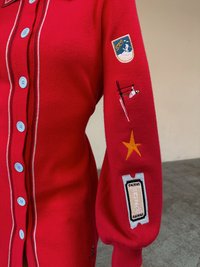 Czerwony dziergany sweter z niebieskimi guzikami, ozdobiony naszywkami: emblematem górskim, narciarzem, gwiazdą oraz wzorem biletu na rękawie.