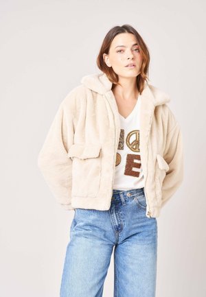 Femme portant une veste beige duveteuse sur une chemise blanche avec des lettres dorées et marron, associée à un jean bleu taille haute sur un fond uni.