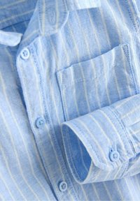 Vaaleansininen puuvillapaita, jossa ohuet valkoiset pystysuorat raidat, etutasku ja siniset napit hihansuissa sekä paidan etukappaleella.