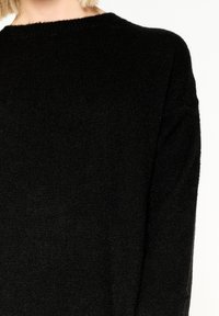 Svart stickad tröja med rund halsringning, långa ärmar och en mjuk, texturerad finish. Utseendet är enkelt och minimalistiskt.