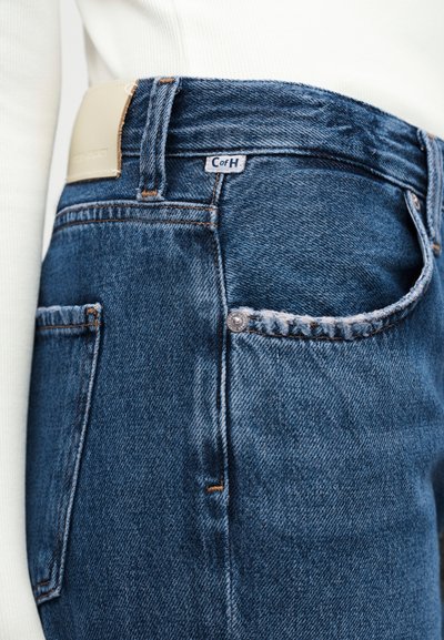 Κοντινό πλάνο τζιν τσέπης από μπλε τζιν με δερμάτινο έμπλαστρο και μικρή ετικέτα "CofH", φορεμένη με λευκό τοπ.