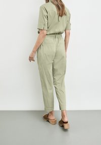 Jasnozielony kombinezon z krótkimi rękawami i elastycznym pasem, z dwoma tylnymi kieszeniami. Noszony z brązowymi sandałami na obcasie z drewna.