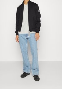 Chaqueta bomber negra con cremallera frontal, puños acanalados y detalle de parche. Lleva una camiseta gris debajo y está combinada con unos jeans acampanados de color azul claro y zapatos negros.