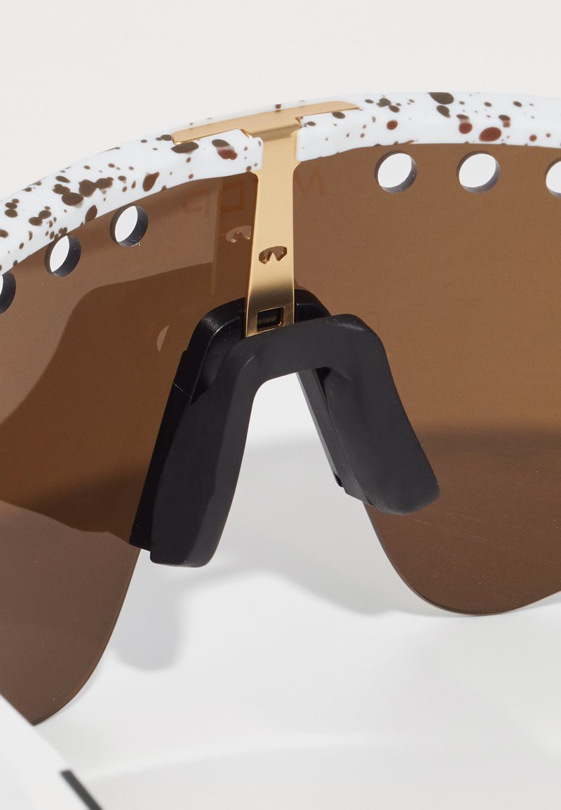 Zonnebril met een wit gevlekt montuur, gouden accenten, bruin getinte glazen en een zwarte neuspad voor comfort. Ontworpen voor functionaliteit en stijl.