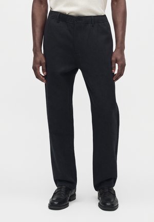 Mann trägt schwarze gerade geschnittene Hosen und schwarze Lederschuhe und steht vor einem schlichten weißen Hintergrund.
