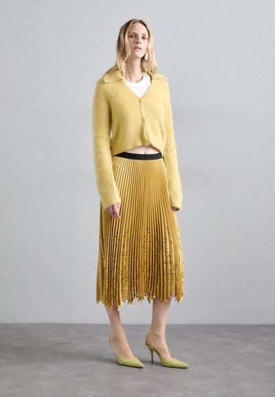 Cardigan galben pufoasă, scurtă, peste un top alb, asortată cu o fustă plisată galbenă cu un model texturat, completată cu tocuri verzi.