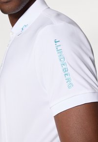 Hvit polo skjorte med en turkise brodert logo på ermet. Laget av et glatt, lett materiale med et klassisk kragedesign.