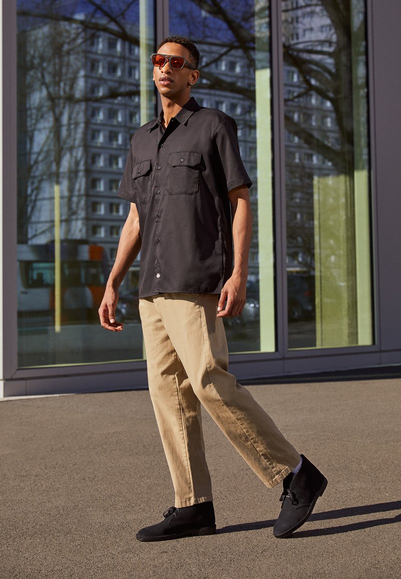 Zwarte kortebloemshirt met twee zakken, beige broek en zwarte schoenen. Reflecterende zonnebril maakt de outfit compleet tegen een moderne glazen achtergrond.