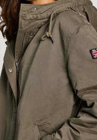 Oliwkowa kurtka z kapturem wykonana z gładkiego materiału. Posiada zasuwaną z przodu, zatrzaski, naszywkę z logo na rękawie oraz regulowany sznurek w kapturze.