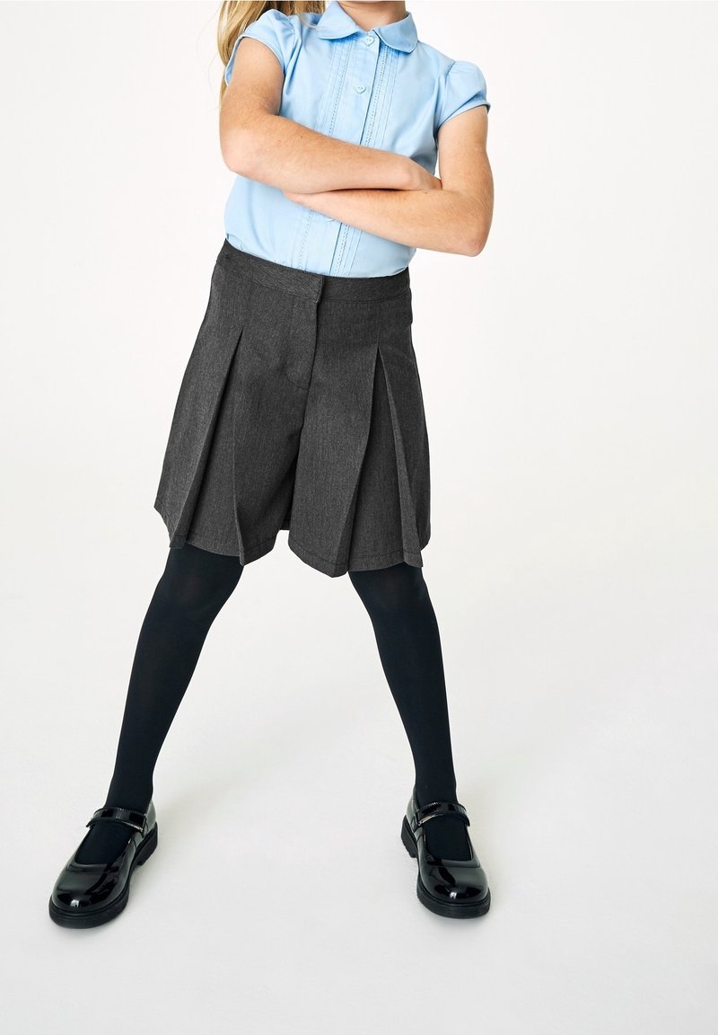Enfant portant une chemise à manches courtes bleu clair, une jupe plissée gris foncé, des collants noirs et des chaussures Mary Jane noires, debout les bras croisés.