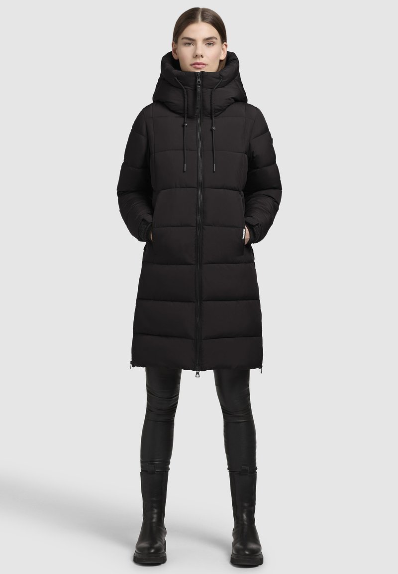 Abrigo largo negro de plumas con cierre de cremallera, cuello alto y capucha con cordón ajustable. Presenta un diseño acolchado y cremalleras laterales.