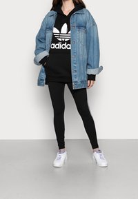 Jeansjacka med uppvikta ärmar, svart hoodie med vit logga, svarta leggings med vita ränder och vita sneakers med lila detaljer.