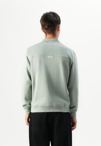 Hombre vestido con sudadera de manga larga gris claro-verde y pantalones negros, de pie con la espalda a la cámara contra un fondo blanco liso.