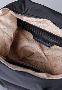 Zwarte schoudertas met een zachte buitenkant, beige stoffen voering met herhaalde logo's en een met rits afsluitbaar binnenvak.