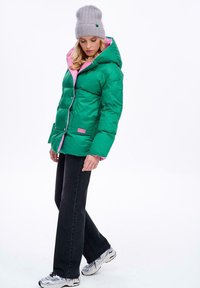 Zielona puchowa kurtka z różową podszewką, wyposażona w kaptur, zatrzaski i boczne kieszenie. W połączeniu z czarnymi jeansami i srebrnymi trampkami.