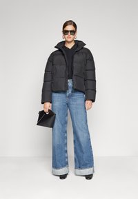 Giacca a vento nera con colletto alto, abbinata a jeans chiari a gamba ampia. Accessorio: piccola borsa nera a tracolla. Modella che indossa occhiali da sole.