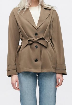 Mujer con una chaqueta taupe con cinturón y botones negros sobre una camiseta blanca y vaqueros azul claro, de pie frente a un fondo liso.