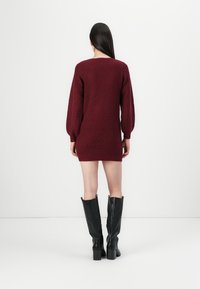 Burgundské pletené šaty s dlhými balónovými rukávmi, priliehavým spodkom a textúrovaným povrchom; vyskúšané s vysokými čiernymi koženými blokovými podpätkami.