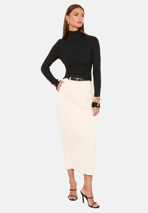 Moteris dėvi juodą ribotą golfą, baltą aukšto juosmens midi sijoną, juodą diržą, auksinius apyrankes ir juodus atvirus aukštakulnius, stovinti.