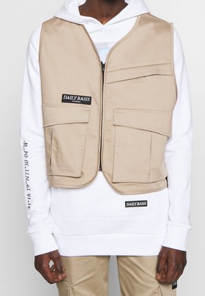 Personne portant un gilet utilitaire beige par-dessus un sweat à capuche blanc avec texte noir sur la poitrine et la manche, et un pantalon beige avec des détails de poche.