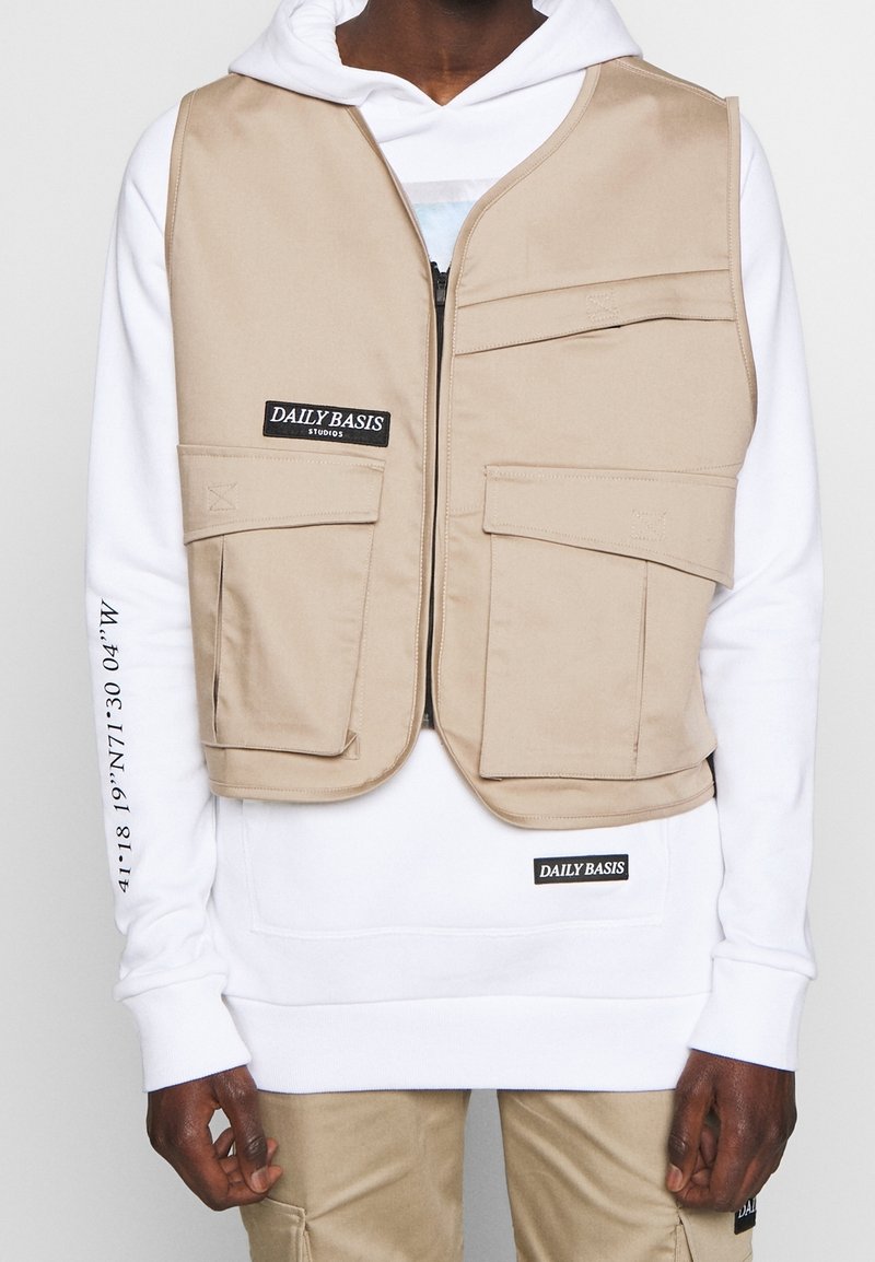 Personne portant un gilet utilitaire beige par-dessus un sweat à capuche blanc avec texte noir sur la poitrine et la manche, et un pantalon beige avec des détails de poche.