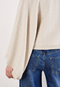 Maglione beige a trama con polsini a coste, caratterizzato da un motivo a maglia morbida. Abbinato a jeans in denim blu, che mettono in mostra i dettagli delle tasche posteriori.