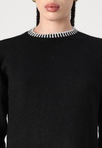 Pull en tricot noir avec une finition texturée, présentant un col côtelé blanc contrastant. Aucun motif visible ni détail matériel.