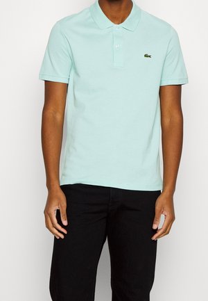 Homme portant un polo bleu clair à manches courtes avec un petit logo crocodile vert sur la poitrine et un pantalon noir.