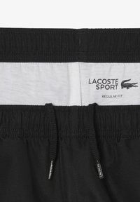 Zwarte sportshorts van elastisch materiaal met een trekkoord en textuur. Het label vermeldt "LACOSTE SPORT" met "REGULAR FIT" op een witte achtergrond.