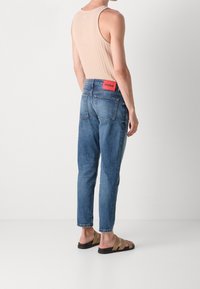 Persona con una camiseta sin mangas color beige claro, jeans de mezclilla azules con una etiqueta roja "HUGO" y sandalias color tostado, de pie contra un fondo blanco liso.