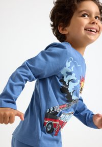 Junger Junge, der lächelt und rennt, in blauen Pyjamas mit einem Feuerwehrwagen, Rauch und Flammenmotiv auf dem Shirt.