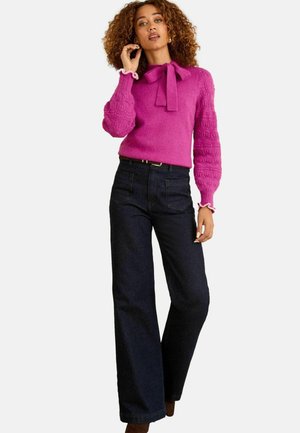 Fuchsia Strickpullover mit einem Bandkragen und strukturierten Ärmeln, kombiniert mit hochgeschnittenen, dunkelblauen Jeans und braunen Stiefeletten mit Absatz.