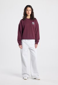 Sweat-shirt bordeaux à épaules tombantes avec un petit patch graphique, associé à un pantalon de survêtement gris clair à jambes larges avec des bandes latérales noires.
