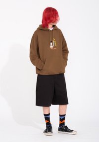 Felpa marrone con grafica frontale, cappuccio con stringa e tasca a marsupio, abbinata a pantaloni corti neri e calze a fantasia, indossata con sneakers nere.