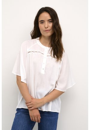 Blouse blanche à manches courtes avec un col rond, présentant des découpes décoratives et une patte de boutons ; portée avec un jean bleu.