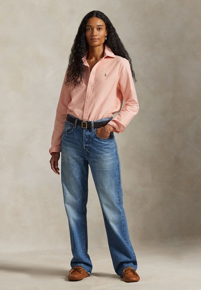 Jeune femme aux longs cheveux bouclés portant une chemise rose clair boutonnée, un jean bleu avec ceinture noire et des mocassins marron.