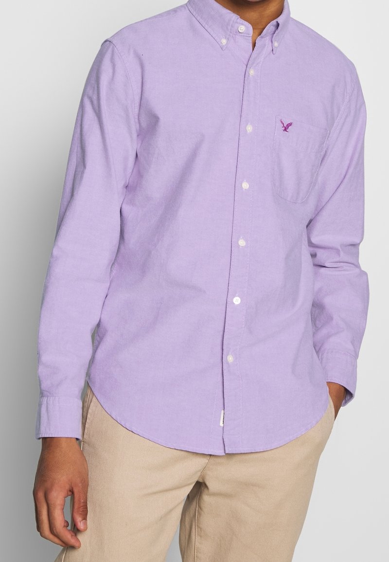 Camicia button-down viola chiaro con taschino sul petto, colletto casual e bottoni bianchi. Il tessuto sembra morbido con un leggero dettaglio di trama.