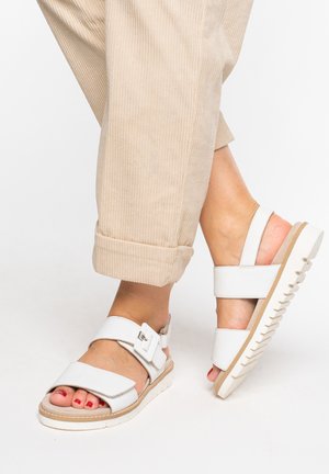 Voeten met witte sandalen met gespen, gecombineerd met beige opgerolde ribfluwelen broeken tegen een effen achtergrond.