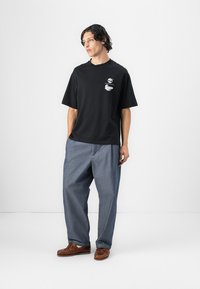 Black short-sleeve t-shirt with a chest graphic, paired with loose blue denim pants and brown leather loafers, set against a plain background.