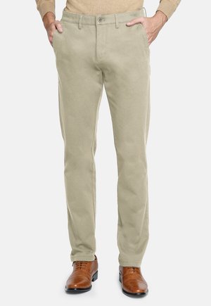 Hombre vestido con pantalones rectos beiges, suéter marrón y zapatos de vestir de cuero marrón, con las manos en los bolsillos.