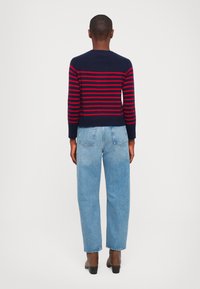 Sini ja punasi raidoilla oleva neule, joka on vyötäröltä tiukka, yhdistettynä vaaleansinisiin korkeavyötäröisiin farkkuihin ja ruskeisiin nilkkureihin. Takapuolen näkymä.