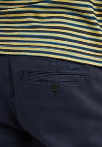 Námořnické bavlněné kalhoty mají zadní kapsu s černým knoflíkem. Částečně viditelná je pruhovaná košile v žluté, černé a modré barvě nahoře.